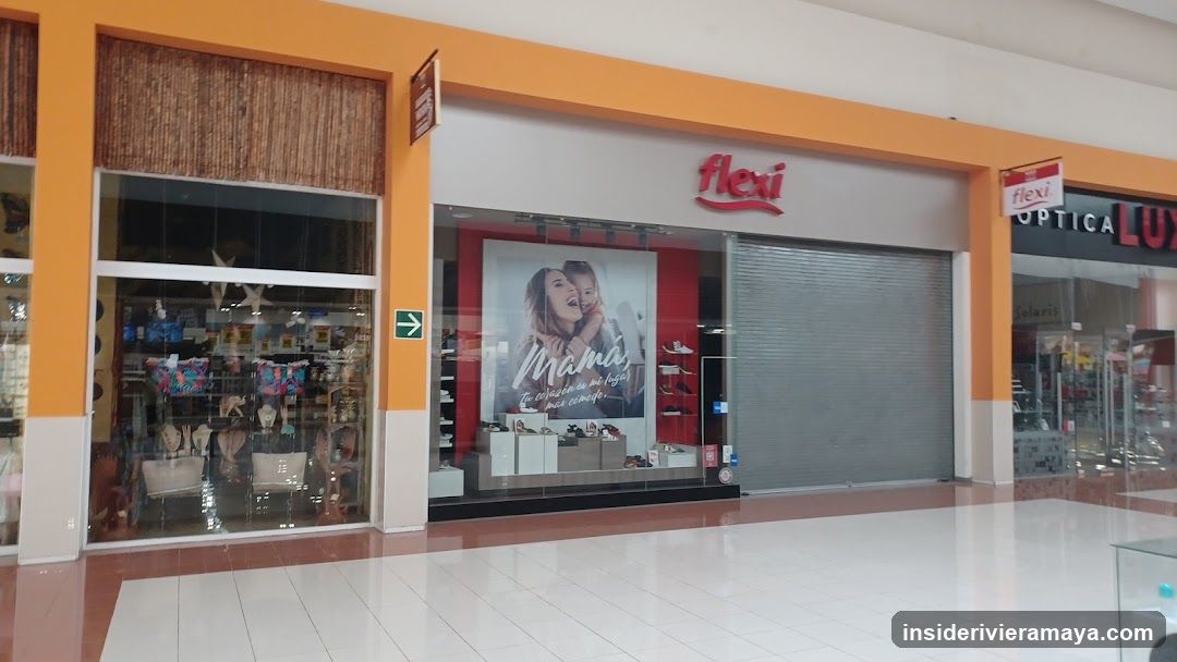 A view of a shopping mall corridor featuring storefronts with orange and white facades. The central store displays a red 'flexi' sign above a large window advertisement featuring a mother and child, alongside a closed metal security shutter. To the left, a store with bamboo blinds displays clothing and accessories. To the right, signage for 'OPTICA LUX' is partially visible.