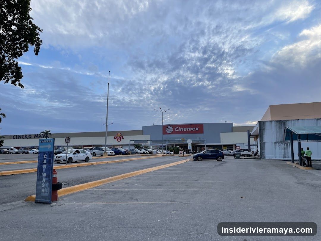 A wide shot of a shopping center parking lot during twilight. The building facade features a large red sign for 'Cinemex' and a smaller sign for 'Dots'. A blue vertical sign in the foreground indicates a 'CAR WASH ENTRANCE'. Several cars are parked in the lot, and a few people are standing near a small structure on the right.