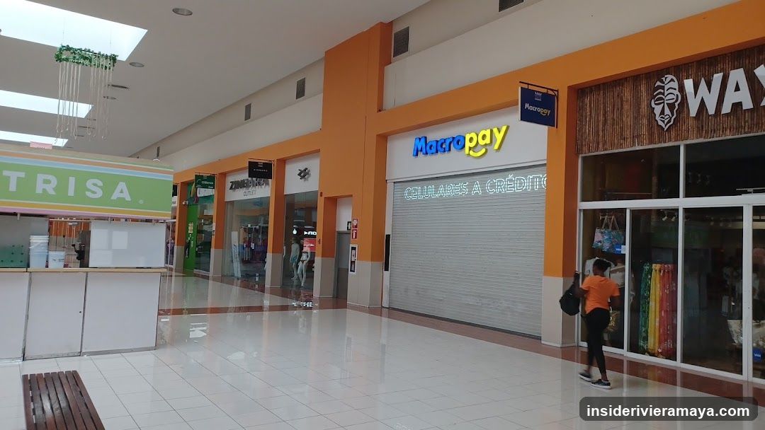 A view of a shopping mall corridor featuring storefronts with orange and white facades. Visible signs include 'TRISA' on a green kiosk, 'Macrogy' above a closed shutter, and 'WAY' with a mask logo on a wooden-textured wall. A person is standing near the glass entrance of the store on the right.