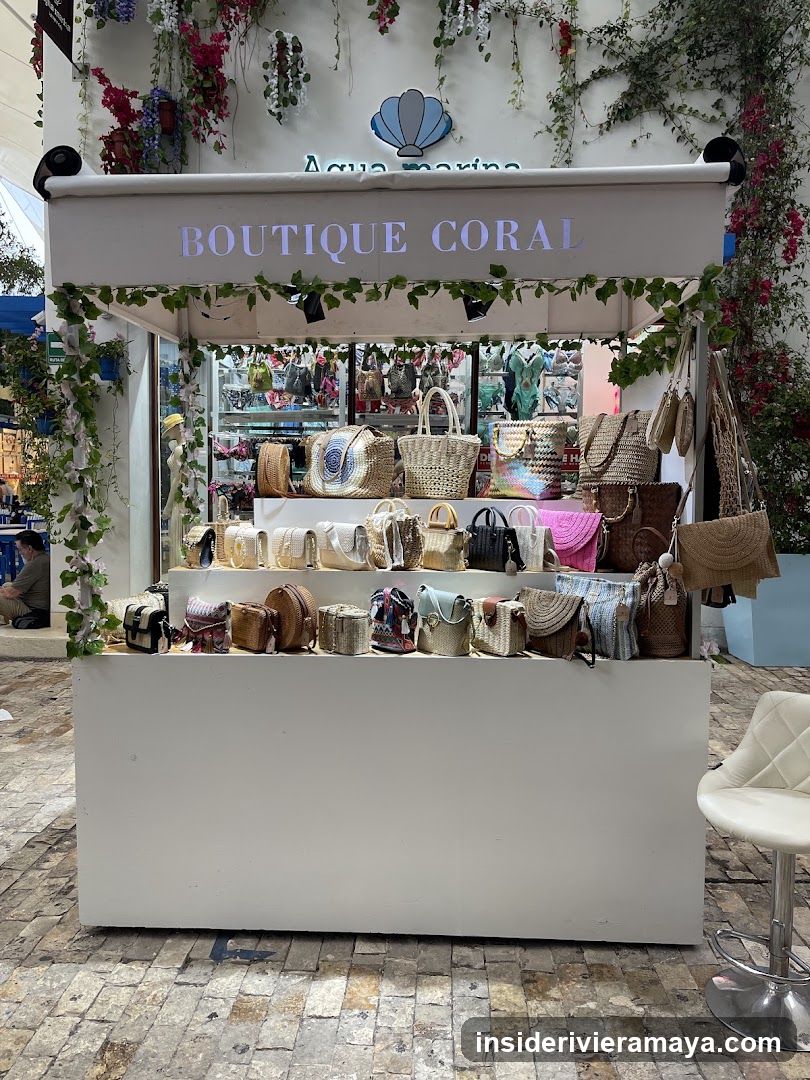 A white outdoor kiosk or boutique stall with a sign reading 'BOUTIQUE CORAL' on the front valance and 'Aqua marina' with a shell logo above it. The display features multiple shelves filled with various handbags, including woven straw totes, leather satchels, and colorful shoulder bags. Greenery vines drape from the top of the structure, and a white stool is visible in the foreground.