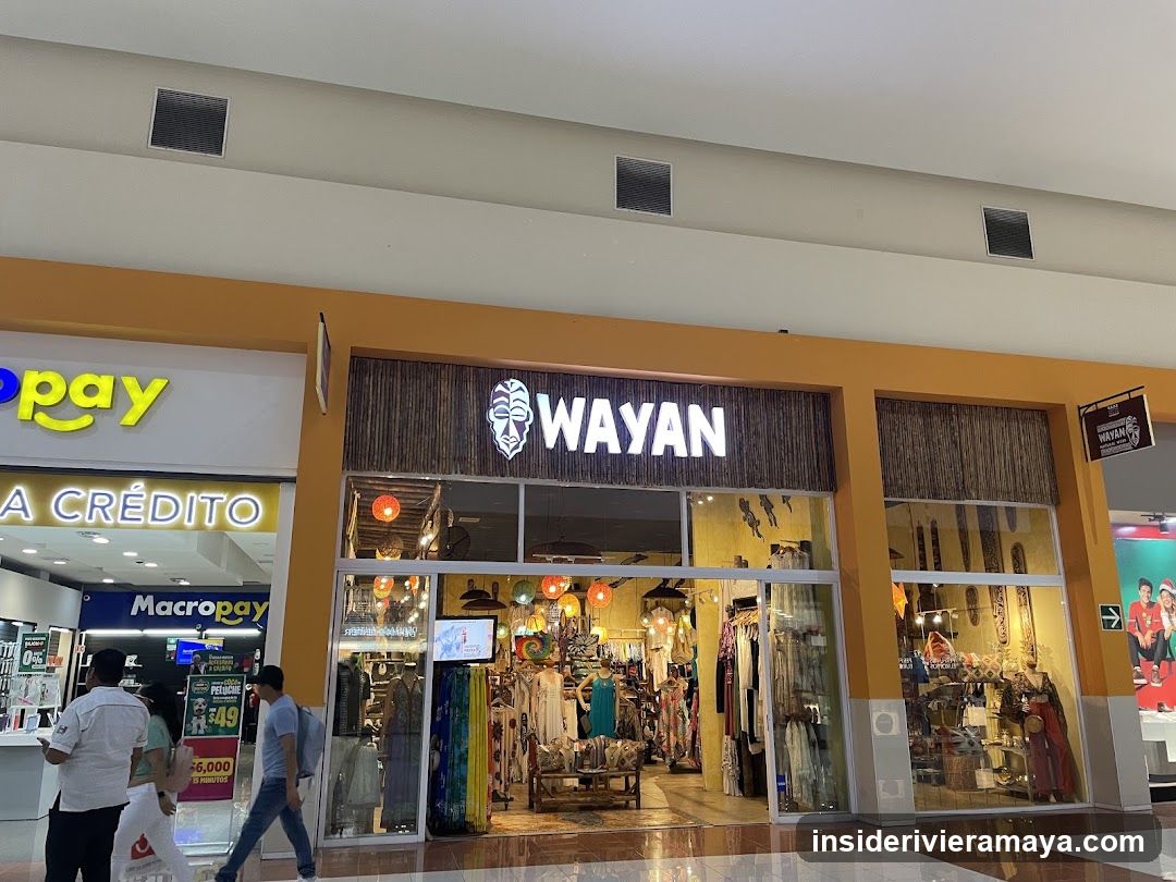 A storefront with a prominent sign reading 'WAYAN' in white letters on a dark, textured background. The store has large glass windows and doors, displaying various clothing items, including dresses and shirts, as well as accessories like bags and hats. Inside, colorful lanterns hang from the ceiling. To the left, a neighboring store named 'MacroPay' is visible, with signage indicating 'A CRÉDITO'.