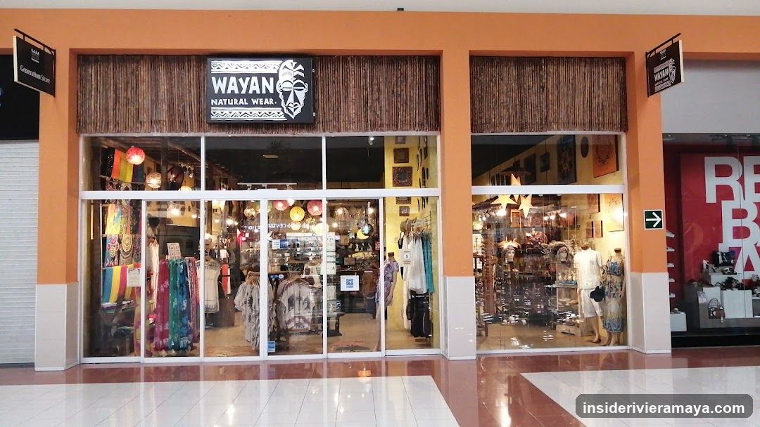 A storefront with an orange facade and large glass windows displaying merchandise. A black sign above the entrance reads 'WAYAN NATURAL WEAR' next to a white mask graphic. The store interior is visible through the glass, showing clothing racks, mannequins, and hanging decorations.