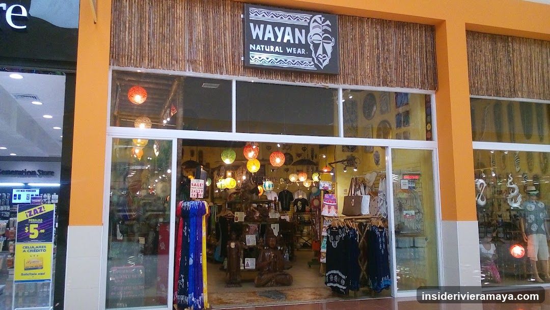 A storefront with orange pillars and bamboo-style wall covering. A black sign reads 'WAYAN NATURAL WEAR' with a tribal mask graphic. The store has large glass windows and doors, revealing hanging lanterns, clothing racks, and wooden statues inside. A yellow poster is visible on the window to the left.