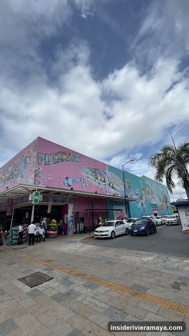 A wide-angle exterior shot of a large commercial building with a colorful mural. The facade is painted pink and teal, featuring the name 'PLAYMART' in large, stylized letters along with illustrations of toys and fish. A green pharmacy cross sign is visible near the entrance. Several cars are parked in front, and people are standing near the entrance on the sidewalk.