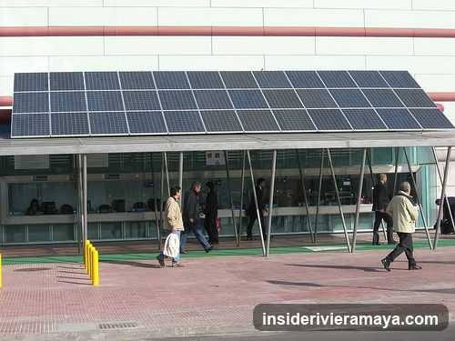 A modern building facade featuring a large, angled canopy covered in solar panels. The structure is supported by angled metal poles. Beneath the canopy, there is a glass-walled service area with several people walking past or standing near the counters. Yellow bollards are visible on the paved walkway in the foreground.