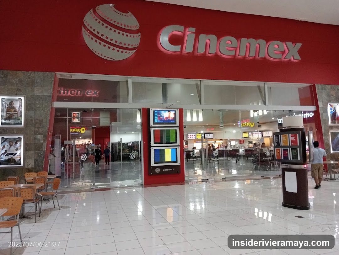 A wide shot of the exterior entrance to a Cinemex movie theater located inside a shopping mall. The facade features a large red sign with the silver 'Cinemex' logo and a film reel graphic. Below, glass doors provide a view into the lobby, where concession stands and seating areas are visible. A digital kiosk displaying movie posters stands in the foreground on the tiled floor.